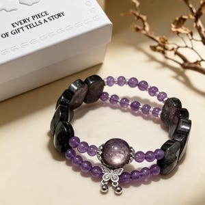 May include: A double-strand bracelet featuring purple amethyst beads and dark gray faceted stones. The bracelet includes a large central purple bead, a silver-toned butterfly charm, and small silver accents. A white box with the text EVERY PIECE OF GIFT TELLS A STORY is visible.