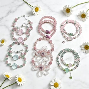 Puede incluir: Una colección de pulseras de cuentas con piedras preciosas rosas y verdes, detalles de cristal transparente y dijes en forma de flor. Las pulseras incluyen varios diseños con cuentas en forma de rosa, colgantes de corazón y flores de cuentas.