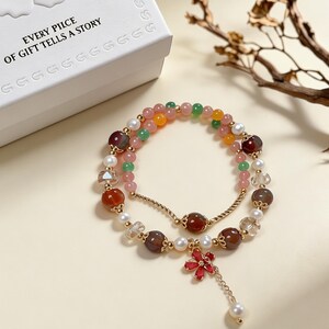 May include: A multi-strand beaded bracelet featuring a mix of round pink, green, yellow, and dark red agate beads, white pearls, and clear faceted crystals. The bracelet includes gold-toned metal spacers and a red flower charm with a dangling pearl.