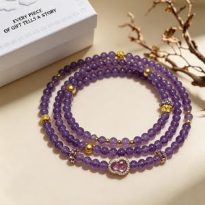 May include: A multi-strand beaded bracelet featuring round purple amethyst beads, interspersed with small gold-toned spherical spacers, textured gold-toned accents, and a central pink stone charm framed by small clear stones. A white box is visible.