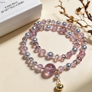 May include: A beaded bracelet featuring alternating translucent pink, clear, and metallic grey beads. A large, central translucent pink bead hangs with a small gold-toned bell charm. The bracelet is positioned near a white box with the text EVERY PIECE OF GIFT TELLS A STORY.