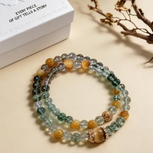 May include: A double-strand beaded bracelet featuring translucent green rutilated quartz beads, opaque golden-yellow tiger eye beads, and two gold-toned metal spacers with clear crystals. The bracelet is presented near a white box with the text EVERY PIECE OF GIFT TELLS A STORY.