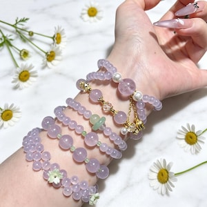 Puede incluir: Una colección de pulseras elásticas superpuestas con cuentas lisas de color morado claro, perlas blancas, cristales transparentes facetados y pequeños dijes de metal dorado. Una pulsera incluye un detalle de flor tallada en verde pálido.