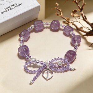 May include: A bracelet featuring polished purple amethyst beads, small white pearls, and translucent purple crystal beads. The bracelet includes a decorative bow made of purple crystal beads and a clear heart-shaped pendant hanging from the center.