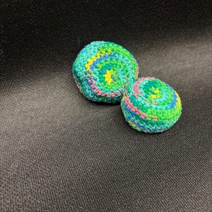 May include: Two handmade crochet balls in shades of green, blue, yellow, and pink. The balls have a spiral design and are set against a dark background. The yarn appears to be cotton.