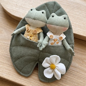 Peut inclure: Deux poupées grenouilles faites à la main dans une poche en forme de feuille verte. L'une porte une combinaison jaune, l'autre une chemise blanche avec broderie florale. Une fleur blanche avec un centre jaune est attachée.