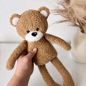 Könnte beinhalten: Ein handgefertigter brauner Teddybär mit strukturiertem, plüschigem Aussehen. Der Bär hat weiße Akzente an Ohren und Schnauze sowie eine schwarze Nase. Der Bär wird von einer Person gehalten.