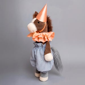 Könnte beinhalten: Ein Plüschpferd-Spielzeug, das für eine Party gekleidet ist. Das Pferd hat einen braunen Körper, weiße Gesichtsmarkierungen und einen grauen Schwanz. Es trägt einen pfirsichfarbenen Partyhut, einen Rüschenkragen und blau-weiß gestreifte Pumphosen. Das Spielzeug steht auf grauem Untergrund.