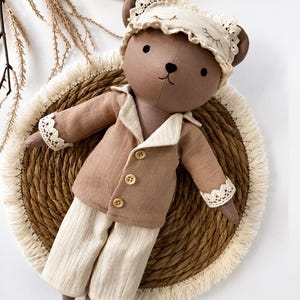 Könnte beinhalten: Eine braune Teddybär-Puppe mit einer beigen Schlafmaske mit Spitzenbesatz und einem passenden Pyjama-Set. Die Pyjama-Jacke hat Holzknöpfe und Spitzensaum an den Ärmeln. Die Puppe liegt auf einer gewebten Matte.