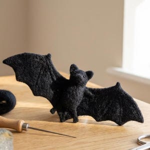 May include: A black needle-felted bat figurine with outstretched wings, positioned next to a wooden-handled felting needle, a small coil of black wool roving, and a pair of silver metal scissors on a light-colored wooden surface.