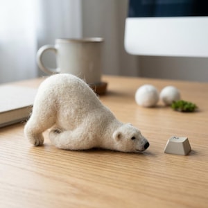 Könnte beinhalten: Eine nadelgefilzte weiße Eisbärenfigur in einer spielerischen Verbeugungspose, platziert auf einer Holzoberfläche neben einer einzelnen beigen Computertastaturtaste mit dem Buchstaben M und zwei kleinen weißen Filzkugeln im Hintergrund.