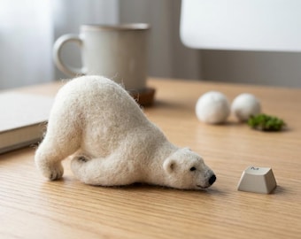 Può includere: Una statuetta di orso polare in lana infeltrita bianca in una posa giocosa di inchino, posizionata su una superficie di legno accanto a un singolo tasto di tastiera beige con la lettera M e due piccole sfere di feltro bianco sullo sfondo.