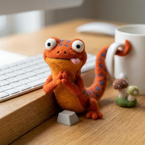 May include: A needle-felted orange lizard figurine with blue spots, large white eyes, and a small pink tongue sticking out. The lizard has its front paws pressed together in a prayer-like gesture and a long, curved tail resting against a white mug.