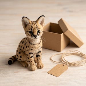 Op de afbeelding: Een naaldvilten serval kitten beeldje met beige vacht en zwarte vlekken, voorzien van grote oren, donkere gezichtsmarkeringen en een lange staart. Naast het beeldje staat een open bruine kartonnen geschenkdoos en een leeg bruin papieren label.