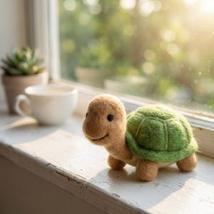 Könnte beinhalten: Eine gefilzte Schildkrötenfigur mit beigem Kopf und Gliedmaßen sowie einem strukturierten grünen Panzer. Die Schildkröte hat kleine schwarze Perlenaugen und einen aufgestickten Mund, gefertigt aus weichen Wollfasern auf einer weißen Fläche.