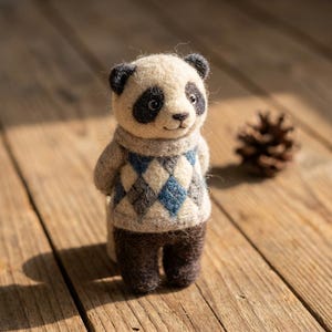 Könnte beinhalten: Eine gefilzte Panda-Figur, die einen beigen Pullover mit blau-grauem Rautenmuster trägt. Der Panda hat schwarze Ohren, schwarze Augenpartien und dunkelbraune Beine. Die Figur ist aus texturierten Wollfasern gefertigt und hat ein kleines Lächeln.