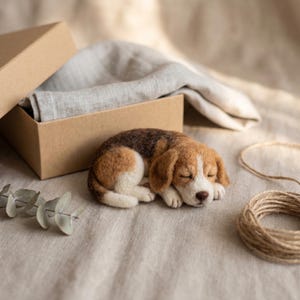 May include: A needle-felted beagle puppy figurine in a sleeping position, featuring a tri-color coat of brown, black, and white wool. The small, handcrafted dog sculpture is positioned next to an open cardboard gift box with a light gray fabric.