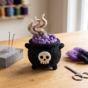 May include: A needle-felted black cauldron featuring a cream-colored skull on the front. The cauldron is filled with various shades of purple felted spheres and topped with swirling, light brown felted smoke rising from the center.