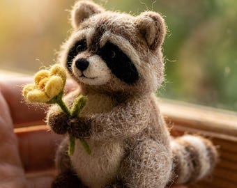 May include: A small needle-felted raccoon figurine crafted from fuzzy brown, white, and black wool. The raccoon has a distinct black mask around its eyes, a striped tail, and is holding a small, bright yellow felted flower with a green stem.