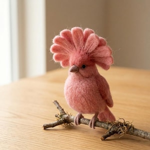 May include: A needle-felted bird figurine in a dusty rose pink color, featuring a prominent, fan-shaped crest on its head, a small dark beak, and black bead eyes. The bird is perched on a natural, textured brown wooden branch with small bits of moss.
