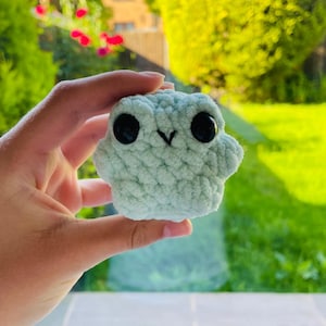 May include: A small, light mint green crochet plush toy with a rounded, textured body. It features two large, shiny black plastic safety eyes and a small, simple black embroidered V-shaped mouth centred between the eyes on its front surface.