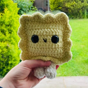 May include: A handmade, crocheted ravioli plush toy. The plush is a light yellow color with a ruffled edge and large black button eyes. It has small, white, crocheted feet. The toy is held in a person's hand.