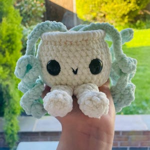 May include: A handmade, plush crochet creature in white with large black eyes and light blue tendrils. The toy has a smiling face and small white feet. The background is a blurred view of greenery.