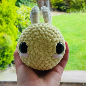 Puede incluir: Un peluche tejido a mano de color amarillo, con grandes ojos negros y pequeñas orejas blancas. El peluche tiene un cuerpo redondo y se sostiene en una mano, con un fondo verde visible.