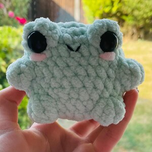 May include: A small, light mint green crochet frog plushie made from soft, textured yarn. The frog features two large, round, glossy black eyes, a small stitched mouth, and two pale pink circular patches on its cheeks below the eyes.