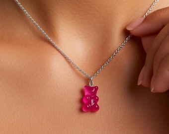 Könnte beinhalten: Eine silberfarbene Metallkettenhalskette mit einem kleinen, durchscheinenden, pinkfarbenen Gummibärchen-Anhänger. Der Anhänger hängt an einer einfachen Gliederkette, die auf der Haut aufliegt und eine glatte, glänzende Oberfläche aufweist.