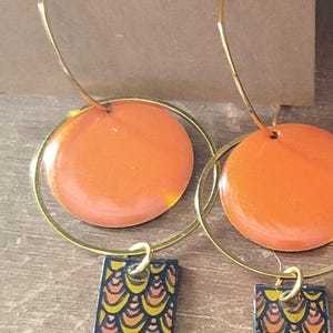 May include: Handmade earrings with orange circular pendants and gold hoops. The earrings have rectangular charms with a colourful scale pattern and yellow teardrop-shaped dangles. The text "Handmade with love" is visible.
