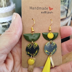Op de afbeelding: Handgemaakte oorbellen met goudkleurige haken en accenten. De oorbellen hebben olijfgroene halve cirkelvormige toppen, cirkelvormige patronen met gele en zwarte bloemmotieven en gele druppel- en rechthoekige vormen. De kaart leest "Handmade with love."