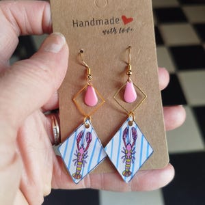Op de afbeelding: Hangende oorbellen met een ruitvormige bedel met een blauw gestreept patroon en een roze en gele kreeftillustratie. Boven de bedel bevindt zich een goudkleurig ruitvormig frame met een roze druppelaccent. De tekst Handmade with love is gedrukt.