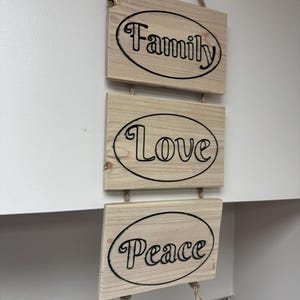 Op de afbeelding: Een houten wanddecoratie met drie rechthoekige platen. Elke plaat heeft een zwart ovaal met de woorden "Family", "Love" en "Peace" in zwarte letters. De platen zijn verbonden met touw.