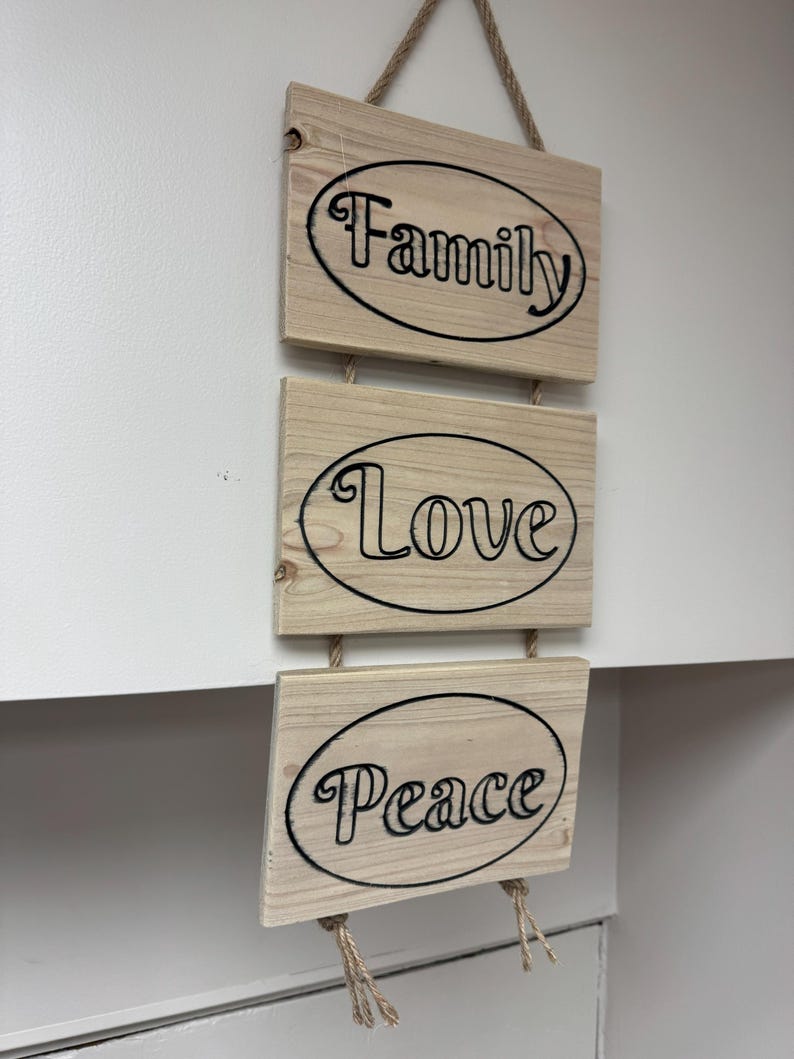 Op de afbeelding: Een houten wanddecoratie met drie rechthoekige platen. Elke plaat heeft een zwart ovaal met de woorden "Family", "Love" en "Peace" in zwarte letters. De platen zijn verbonden met touw.