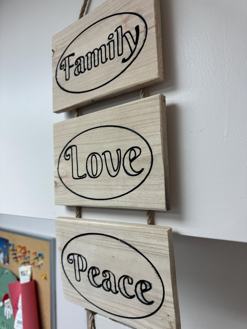 Op de afbeelding: Een houten wanddecoratie met drie rechthoekige platen. Elke plaat heeft een zwart ovaal met een woord in zwart schrift: "Family", "Love" en "Peace". De platen zijn verbonden door een natuurlijk touw.