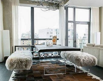 May include: A modern dining room with a large, black, rectangular table with a glass top and four clear acrylic legs. Two white fur benches are placed on either side of the table. A large, branch-like chandelier hangs from the ceiling. A cowhide rug with black and white patches covers the floor.