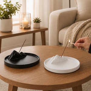 Puede incluir: Dos porta inciensos, uno negro y otro blanco, cada uno con un diseño de montaña. Un palito de incienso está encendido en el soporte blanco. Los soportes están sobre una mesa redonda de madera. Una vela encendida y plantas están en el fondo.