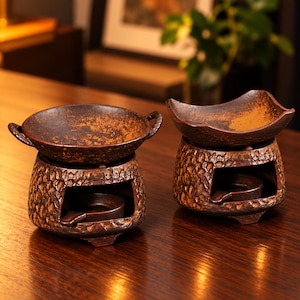 May include: Two dark brown ceramic tea warmers with a textured, rugged exterior finish. Each warmer features a hollow base for a candle and a removable top bowl with small side handles, showcasing a mottled, weathered appearance.