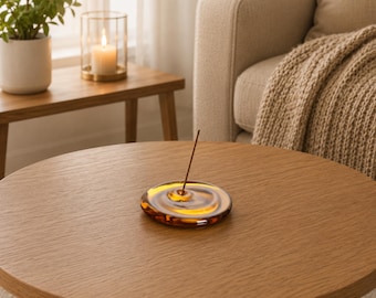 May include: A circular, amber-coloured glass incense holder with a smooth, rippled surface that mimics the appearance of a water droplet. A single, thin, brown incense stick is inserted into the small, centred hole of the glass dish.
