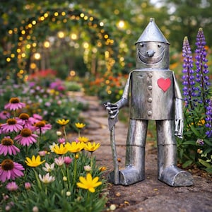 Könnte beinhalten: Eine Metallskulptur des Blechmanns aus Der Zauberer von Oz, mit einem roten Herzen und einem Wanderstock, steht auf einem Gartenweg, umgeben von bunten Blumen. Die Skulptur hat einen konischen Hut und ein lächelndes Gesicht.