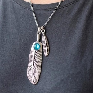 Puede incluir: Collar plateado con colgante de pluma. El colgante presenta una pluma grande y detallada y una pluma más pequeña. Una piedra redonda de color turquesa está engastada en la pluma más grande. El collar se lleva sobre una camiseta gris oscuro.