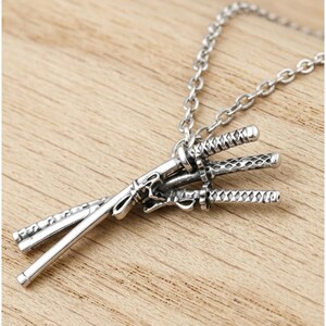 May include: A silver-colored necklace featuring a pendant of three miniature katana swords. The swords are intricately detailed and connected to a chain. The pendant rests on a wooden surface.