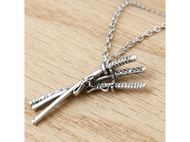 May include: A silver-colored necklace featuring a pendant of three miniature katana swords. The swords are intricately detailed and connected to a chain. The pendant rests on a wooden surface.