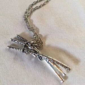 May include: Silver-toned pendant necklace featuring three miniature katana swords. The swords are intricately detailed and tied together, suspended from a delicate chain. The necklace rests on a white fabric surface.