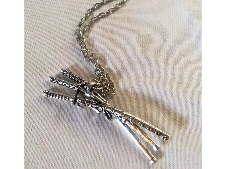 May include: Silver-toned pendant necklace featuring three miniature katana swords. The swords are intricately detailed and tied together, suspended from a delicate chain. The necklace rests on a white fabric surface.