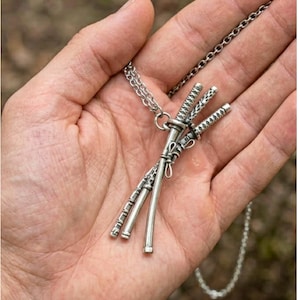 May include: Silver-toned necklace featuring a pendant of three miniature katana swords. The swords are intricately detailed and arranged in a crossed formation, suspended from a delicate chain. The necklace is displayed against a natural background.