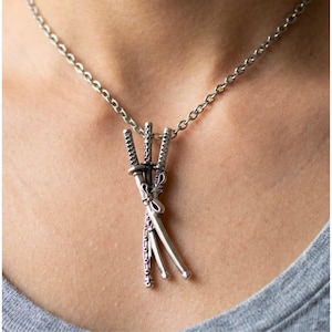 May include: Silver-toned necklace featuring a pendant of three miniature katana swords. The swords are intricately detailed and bound together, suspended from a delicate chain. The necklace is worn against a gray shirt.