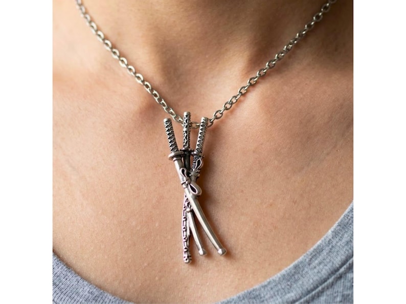 May include: Silver-toned necklace featuring a pendant of three miniature katana swords. The swords are intricately detailed and bound together, suspended from a delicate chain. The necklace is worn against a gray shirt.