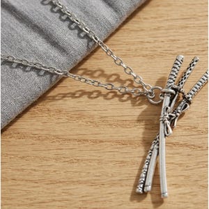 May include: Silver-toned necklace featuring a pendant of three miniature katana swords. The swords are intricately detailed and tied together with a decorative element. The chain is a simple link style, resting on a gray fabric and wooden surface.